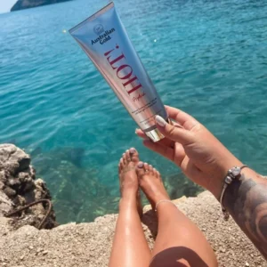 Australian Gold Hot! Tanning Lotion being held out towards the sea, with a woman's tanned legs in the foreground.
